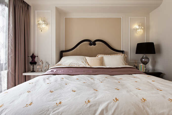 卧室中放置着清晰线条的简约大床，搭配上有金色小花样的白色布艺床品，很是柔软大方。床头的背景墙以米色的乳胶漆刷至而成，在加上白色的镶边，整个的丰富多彩。两边水晶吊灯的设计，一种柔和中的浪漫之感。