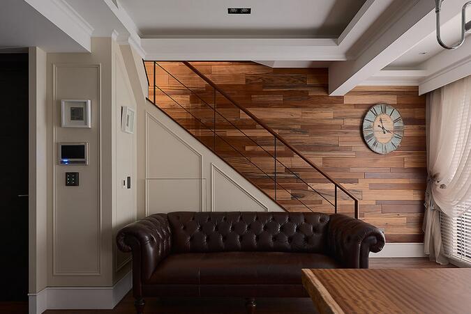 入门处，通往二楼的梯间运用不同原木的纹路铺贴出自然感，楼梯下方的壁面贴覆古典线板，再摆设一张复古拉扣沙发，凸显空间的层次重点。