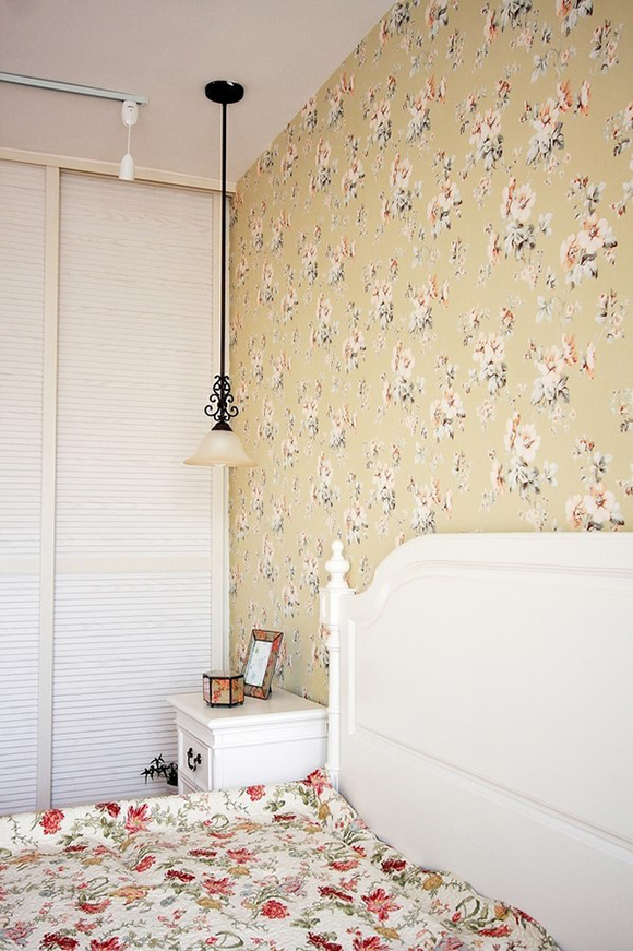 卧室的床品与壁纸诠释了乡村田园的主打元素，精致的吊灯，白漆床头柜上摆放着风格一致的小相框和收纳盒。